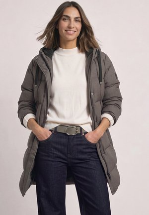 Une femme souriante porte un manteau matelassé gris, un pull blanc, un jean foncé et une ceinture sur un fond clair et uni.