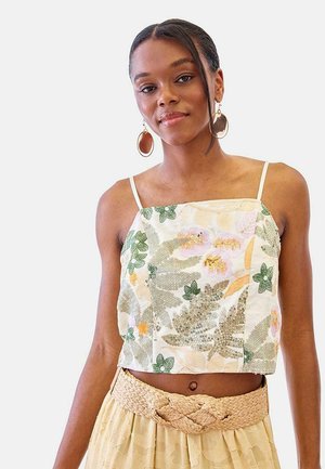Joven con pendientes trenzados lleva un top corto floral con lentejuelas y una falda beige con un cinturón ancho trenzado.