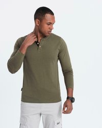 Man i olivgrön långärmad henley tröja och vita cargobyxor, justerar kragen på tröjan, bär en svart armbandsur, mot en enfärgad bakgrund.