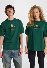 Zielone bawełniane t-shirty z centralnym graficznym logo, krótkie rękawy, luźny krój i okrągły dekolt, noszone z różnymi stylami spodni.