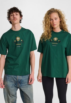 Grüne Baumwoll-T-Shirts mit einem zentralen Logo-Design, kurzen Ärmeln, lockerer Passform und Rundhalsausschnitt, kombiniert mit verschiedenen Hosenstilen.