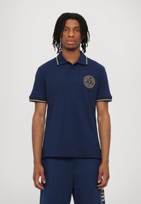 Marineblå polo med gule detaljer, korte ermer og krage. Har en rund logo på brystet. Laget av et glatt stoff.