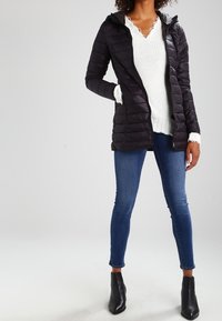 Svart puffjacka med huva, quiltat mönster, vit stickad tröja med spetsdetaljer, blå jeans och svarta ankelstövlar.