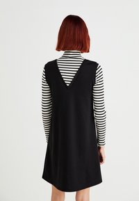 Robe noire sans manches avec un dos en V portée par-dessus un haut ajusté noir et blanc à rayures aux manches longues, tissu lisse et jupe évasée.