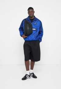 Blå vindjakke med sorte accenter, der har et geometrisk mønster og frontlogo, parret med sorte shorts og sorte sneakers med hvide detaljer.