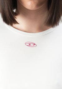 Gros plan d'une personne aux cheveux bruns mi-longs portant un haut blanc avec un petit logo rouge brodé près de l'encolure.