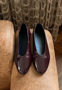 Babies en cuir verni bourgogne avec un bout arrondi et une texture lisse, posées sur une surface brune texturée. Pas de quincaillerie ou d'accents visibles.
