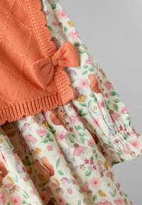 Corpetto lavorato a maglia arancione con dettaglio a fiocco sopra una gonna di cotone a fantasia floreale che presenta animali, fiori e ghiande su uno sfondo crema.