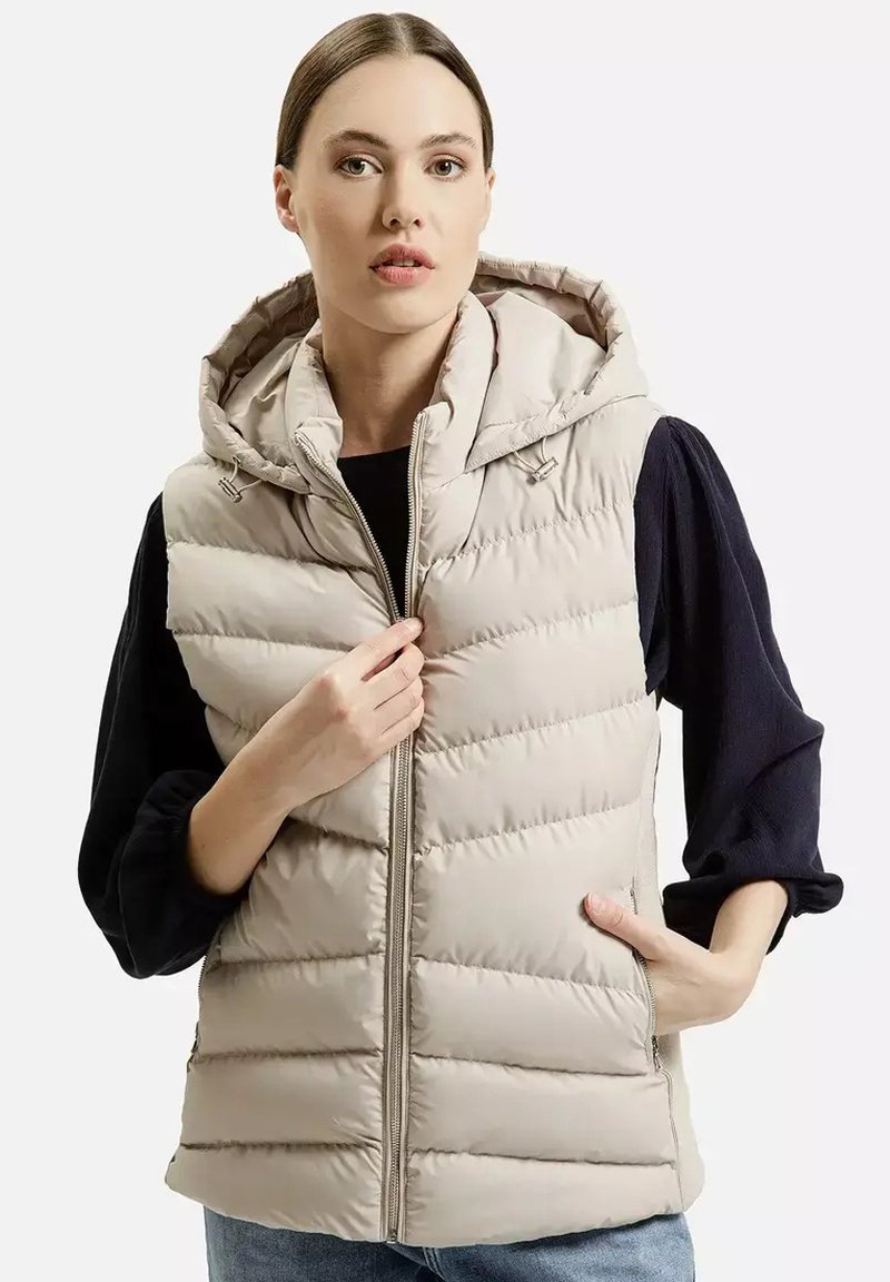 Beżowa kamizelka puchowa z kapturem, z poziomymi przeszyciami, z przodu na zamek błyskawiczny i kieszeniami po bokach. Nosi ją osoba w czarnej bluzie z długim rękawem.
