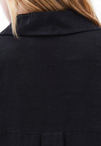 Czarna lniana koszula z złożonym kołnierzykiem i detalem w postaci zakładki na plecach, prezentująca gładką, teksturowaną powierzchnię materiału.