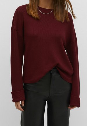 Femme portant un pull côtelé bordeaux avec des poignets retroussés et un pantalon en cuir noir, vue du cou aux hanches sur un fond uni.