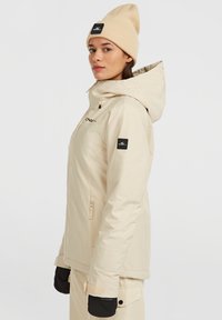 Beige geïsoleerde jas met capuchon, voorzien van een rits aan de voorkant, zijzakken en een logo patch op de mouw. Draagt een beige gebreide beanie.