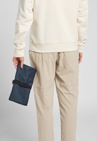 Rollenbeutel aus dunkelblauem Stoff, gesichert mit einem schwarzen Riemen, gehalten in einer Hand, kombiniert mit hellbeigen Hosen und einem cremigen Sweatshirt.