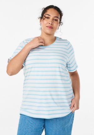 Ženska z kodrastimi lasmi, oblečena v svetlomodro in belo črtasto majico ter modre kavbojke, stoji pred enobarvnim ozadjim.