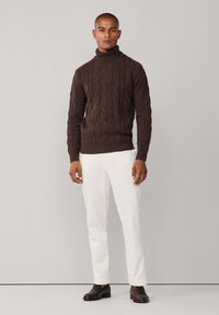 Suéter de cuello alto de punto trenzado marrón combinado con pantalones blancos. El suéter presenta un dobladillo acanalado y un patrón texturizado en toda la prenda.