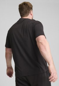 Czarna koszulka z krótkim rękawem, wykonana z gładkiej bawełny, charakteryzująca się luźnym krojem i prostym dekoltem, noszona przez osobę od tyłu.