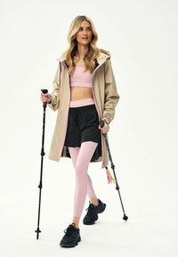 Kobieta w beżowej kurtce, różowym sportowym biustonoszu i legginsach oraz czarnych szortach, idąca z kijkami trekkingowymi na białym tle.