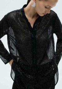 Sort gennemsigtig bluse med palietter, lange ærmer, knaplukning fortil og sidelommer. Stoffet har en tekstureret, glitrende overflade.