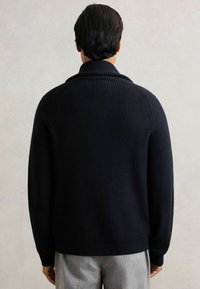 Hombre vestido con un suéter de punto acanalado azul marino oscuro y pantalones gris claro, de pie de espaldas contra un fondo claro y liso.