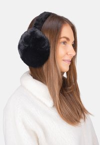 Cache-oreilles en fausse fourrure noire au design arrondi, avec une texture moelleuse et un ajustement confortable autour des oreilles. Porté avec une veste blanche.