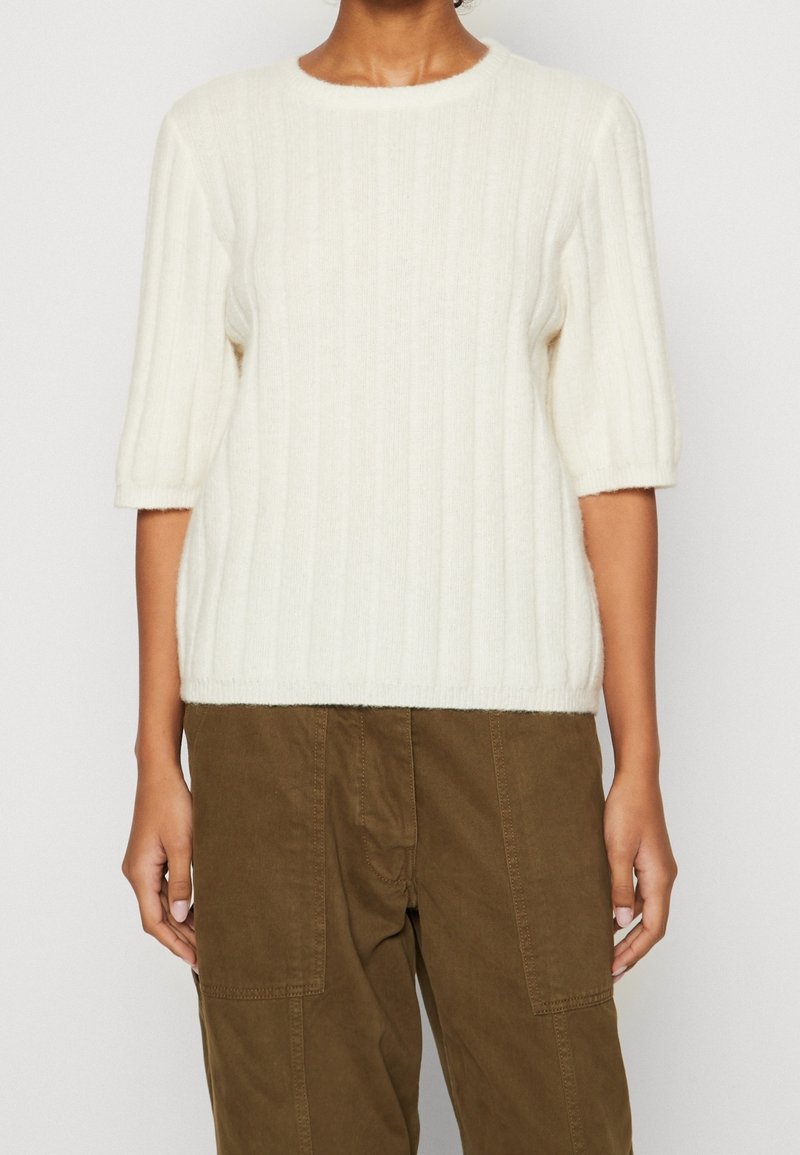 Femme portant un pull blanc côtelé à manches courtes et un pantalon marron avec poches plaquées devant, debout devant un fond uni.