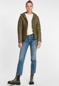 Olivengrønn puffjakke med horisontale quiltede mønstre, frontglidelås og hette. Bært over en kremfarget strikket genser og blå jeans, sammen med ankelstøvler.