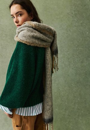 O tânără îmbrăcată cu o pulover tricotat verde, o cămașă cu dungi, pantaloni maro și o eșarfă mare bej, pe un fond verde estompat.