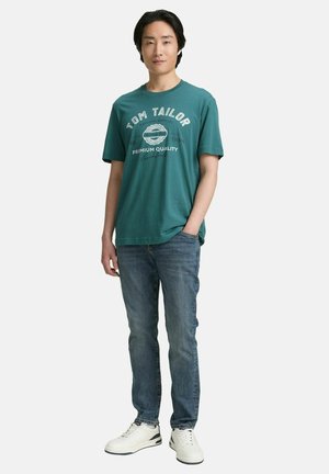 Teal cotton t-shirt featuring white text with logos, paired with blue denim jeans and white sneakers, standing against a plain background.