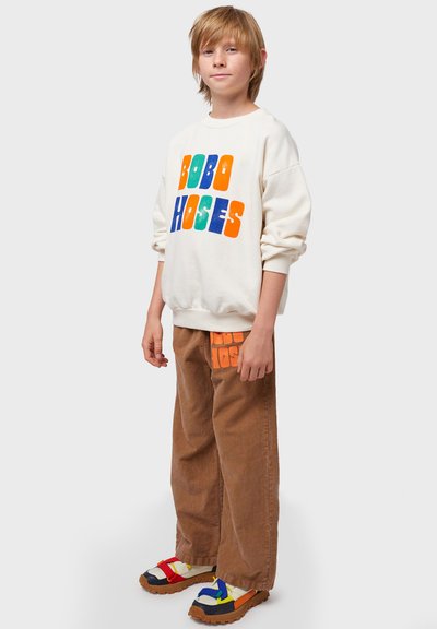 Sudadera crema con impresión colorida "BOBO HOGES," combinada con pantalones de corduroy marrón que muestran un logotipo más pequeño. Sandalias gruesas multicolores.
