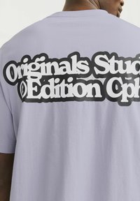 Světle fialové tričko s výrazným černým textem "Originals Studio Edition Cph" na zadní straně, s hladkou, měkkou texturou.