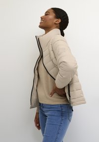 Beige pufferjack met quilted textuur, hoge kraag en zwarte rits. Draagt over een lichtbeige trui en gecombineerd met blauwe jeans.