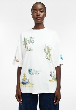 Kvinde iført en hvid oversized t-shirt med akvarelmaling af kaktus, palme, fontæne og skyer, kombineret med mørkeblå jeans.