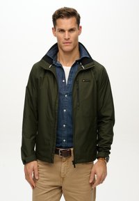 Chaqueta de cremallera verde oliva con un acabado texturizado, con un cuello negro y un pequeño bolsillo en el pecho, combinada con una camisa de denim.
