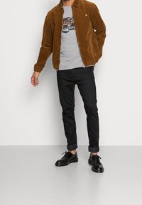 Man wearing brown corduroy jacket, graphic grey t-shirt, black rolled-up jeans, and black leather shoes, standing against white background.