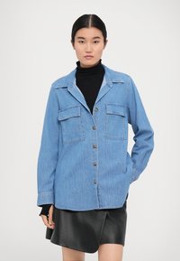 Hellblaues Denimhemd mit zwei Brusttaschen, Knopfleiste und klassischem Kragen, kombiniert mit einem schwarzen Rollkragenpullover und einem Lederrock.