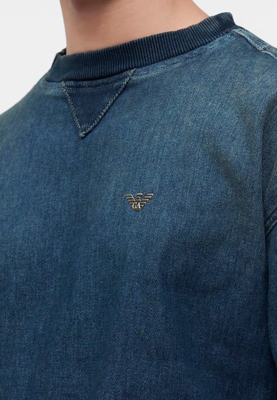 Homme portant un sweat bleu aspect denim avec col côtelé et petit logo métallique sur la poitrine.