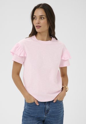 Femme portant un haut rose pâle à manches courtes avec des manches volantées et un jean bleu, les mains dans les poches, regardant sur le côté devant un fond uni.