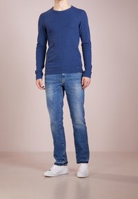 Blå långärmad tröja i stickad tyg, kombinerad med ljusblå jeans och vita sneakers, mot en enkel bakgrund.