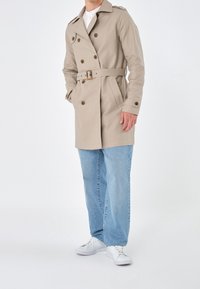 Cappotto beige con design doppiopetto, vita con cintura e bottoni grandi, abbinato a jeans azzurri chiari e sneakers bianche.