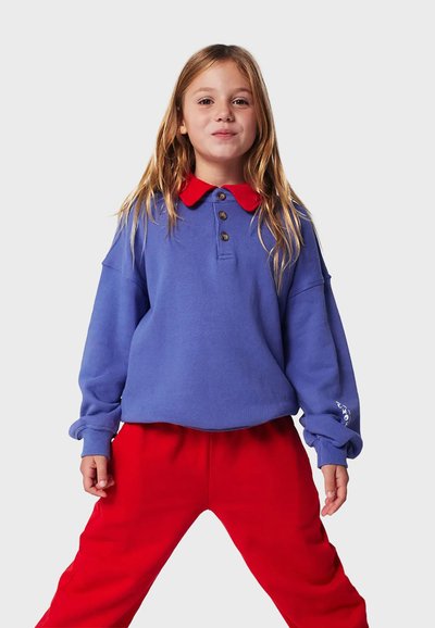 Blaues Sweatshirt mit rotem Kragen und drei Knöpfen, kombiniert mit roten Sweatpants. Besitzt eine leichte Textur und einen lockeren Schnitt mit langen Ärmeln.