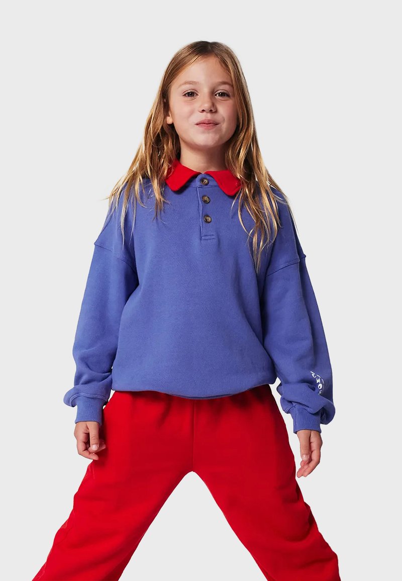 Lila Sweatshirt mit rotem Kragen, ausgestattet mit drei braunen Knöpfen und lockeren Ärmeln, kombiniert mit roten Jogginghosen.