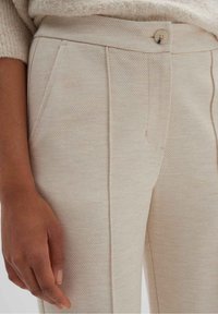 Pantalones de corte beige hechos de tejido texturizado. Presentan bolsillos laterales, un cierre con botón visible y costuras estructuradas. Primer plano del detalle de la tela.