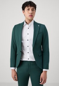 Blazer teal et pantalon assorti en tissu lisse. Chemise blanche avec boutons noirs, présentant un poche carrée et un design épuré.