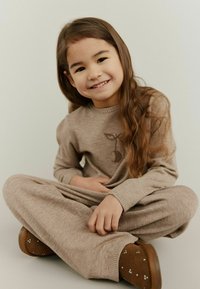 Beige gebreide outfit met lange mouwen en broek, met een eenvoudig dierontwerp op de borst. Bruine instapschoenen met kleine accenten.