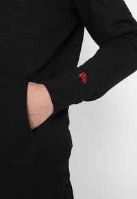 Sudadera negra de corte relajado, con puños y dobladillo acanalados, que presenta un detalle del logotipo rojo en el puño izquierdo. Bolsillo frontal visible.