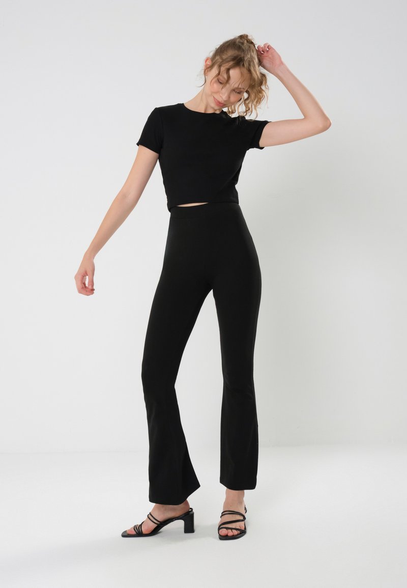 Camiseta negra corta y pantalones acampanados negros de cintura alta, ambos en una tela suave. La modelo los combina con tacones negros con tiras. Diseño simple.