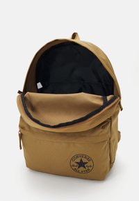 Zaino kaki con un compartimento principale chiuso da zip e una tasca frontale. Presenta una fodera nera e un logo circolare Converse sulla parte anteriore.
