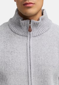 Grå zip-sweater med ribs og tekstureret stof. Metal lynlås med brun lædertræk. Tætsiddende design, ingen synlige mønstre.