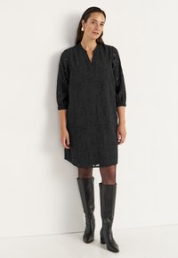 Schwarzes, knielanges Kleid mit einem dezenten texturierten Muster, halblangen Ärmeln und einem V-Ausschnitt, kombiniert mit kniehohen schwarzen Stiefeln.