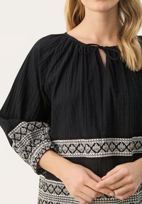 Zwarte blouse met een gestructureerde afwerking, voorzien van een striksluiting bij de hals en een patronen van witte borduursels op de mouwen en zoom.